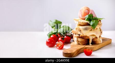 Stockpile of toasted melted cheese pressed sandwiches with ham meat, cherry tomatoes and basil leaves on wooden cutting board on white marble table. B Stock Photo