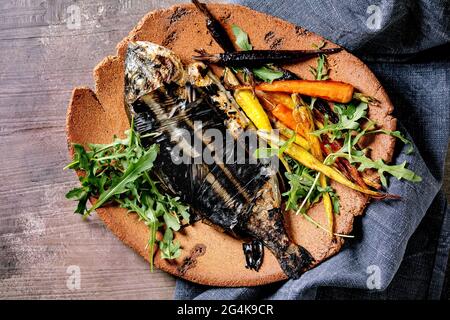 Grilled cooked fresh gutted sea bream or dorado fish on ceramic plate wrapped in bamboo leaves served with herbs, colorful carrots, blue napkin over d Stock Photo