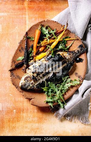 Grilled cooked fresh gutted sea bream or dorado fish on ceramic plate wrapped in bamboo leaves served with herbs, colorful carrots, white napkin over Stock Photo