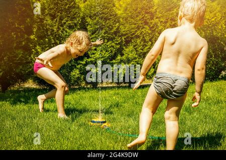 siblings playing with lawn water sprinkler in backyard on hot summer day Stock Photo