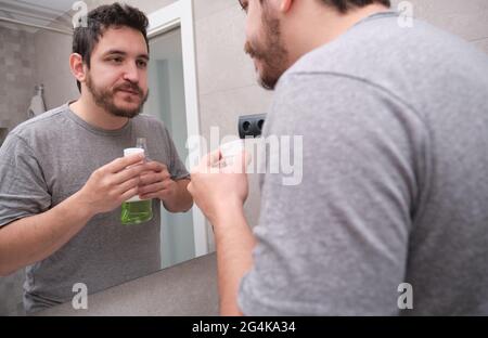 Tired sleepy man rinsing mouth with green mouthwash in bathroom. Teeth care concept. Stock Photo