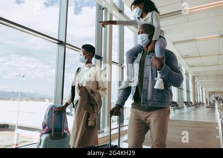 Kid girl on shoulder of man showing airplanes while walking at airport. Family wearing face masks going for boarding airplane. Stock Photo