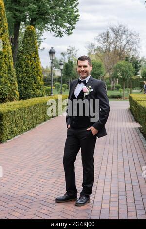 Smiling groom holding hands in pockets on walkway in park Stock Photo