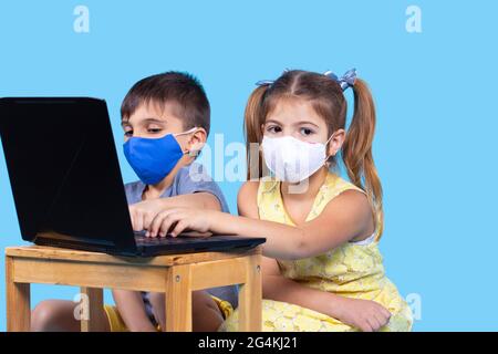 Boy and a girl on the online education. In a protective mask are siting together working with a notebook on a blue background. Stock Photo