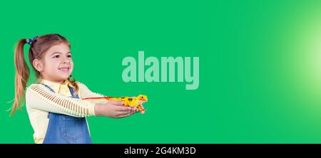 Banner, long format. A schoolgirl shows a toy, lizard. Smiling and keeping the toy in her hands. Green background. Stock Photo