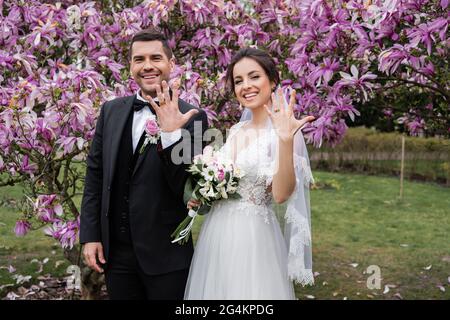 Smiling bride and groom showing rings near magnolia trees Stock Photo