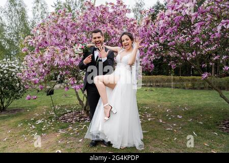 Cheerful newlyweds showing rings near magnolia trees in park Stock Photo