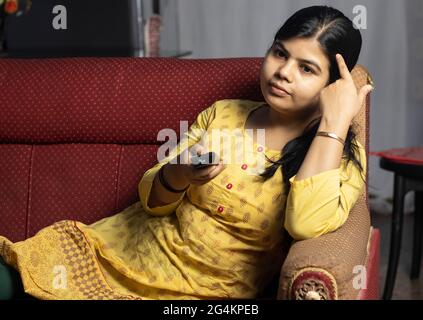 An Indian housewife woman watching television holding remote in hand, sitting on sofa Stock Photo