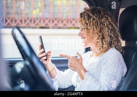 Happy lady speaking in video call conference sit down inside the car on passenger seat holding a phone and smiling to friends - online connection inte Stock Photo