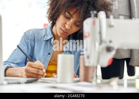 Concentrated African-American dressmaker draws in sketchbook sitting at workplace with modern sewing machine Stock Photo