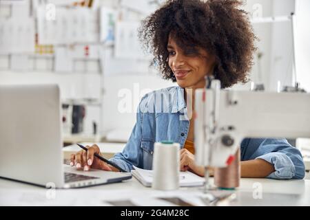 African-American woman uses laptop at workplace with sewing machine Stock Photo