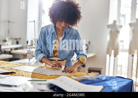Professional African-American dressmaker draws pattern on fabric working at cutting table Stock Photo