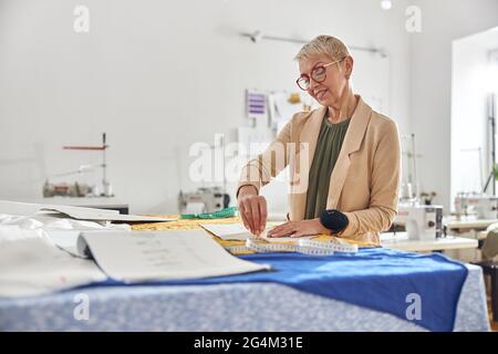 Mature woman designer draws pattern on color fabric at large cutting table in workshop Stock Photo