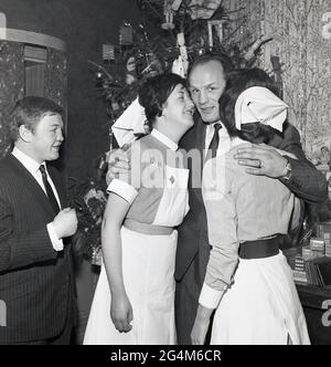 1960s, Famous British heavyweight boxing champion Henry Cooper gives a rueful smile as he hugs two nurses at a christmas reception for the borough's nursing profession.  Former Olympic Flyweight gold medalist and British Featherwight Champion, Terry Spinks looks on in mock jest, Lewisham, Southeast London, England, UK. Henry Copper became a famous figure in British sport after knocking the Legendary American boxer Muhammed Ali to the canvas and was one of the first to capitalise on the growth of the sports celebrity industry, becoming a household name promoting major consumer brands. Stock Photo