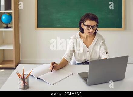 School teacher sitting at desk with laptop and having online class with students Stock Photo