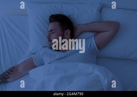 Young man sleeping peacefully at night, lying with closed eyes in comfortable bed in bedroom at home. Stock Photo