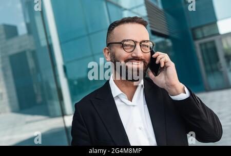 Cheerful middle-aged employee talking on smartphone and smiling while walking near office center, free space Stock Photo