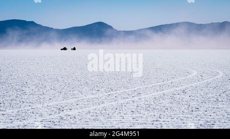 The uyuni salt flat with the mountains, and the silhouettes of two off-road cars in a fog of salt dust in the distance. The tracks of the vehicles dra Stock Photo