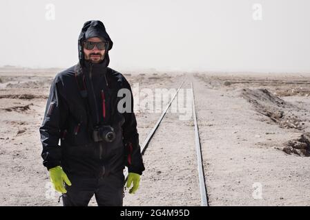A young man wearing a hood, gloves and sunglasses stands with his camera next to some old train tracks in Uyuni, bolivia. Stock Photo