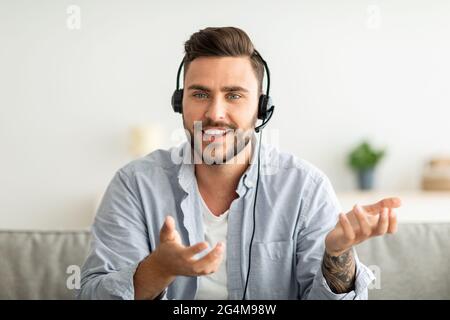 Excited man in headset talking via webcam, having video call with distant colleague, sitting on sofa at home. Stock Photo