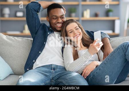 Portrait Of Relaxed Young Multiracial Couple Resting With Smartphone On Couch Stock Photo