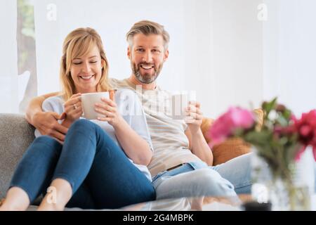 Shot of happy woman and man wearing casual clothes while relaxing on the couch and drinking tea. Stock Photo