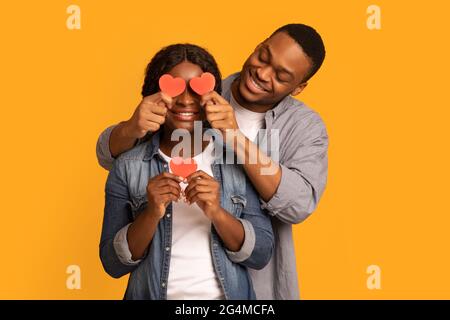Romantic African Guy Covering Eyes Of His Girlfriend With Red Paper Hearts Stock Photo