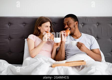 Affectionate multiracial couple having breakfast in bed, eating fresh croissants wth coffee at home Stock Photo