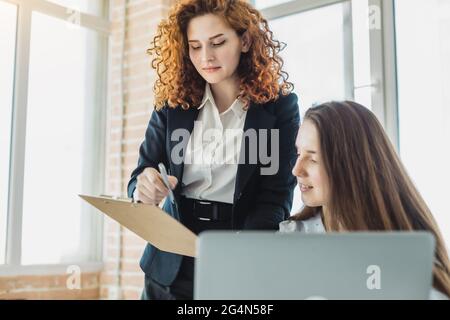 Two beautiful young woman talking and discussing working moments with laptop in the office. Stock Photo