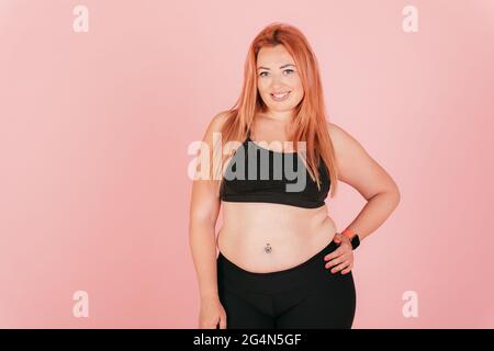 Beautiful happy plus size young woman wearing sporty outfit standing on pink backdrop. Stock Photo