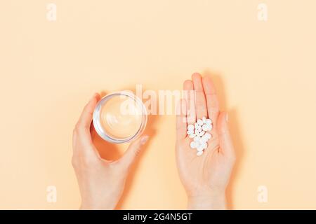 Top view of female hands holding handful medical pills and glass of water on beige background. Stock Photo
