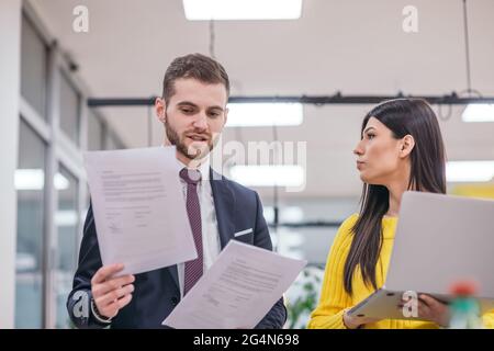 Businessman boss checking the paperwork handed over bus his female employee, while standing in a modern startup office. Stock Photo