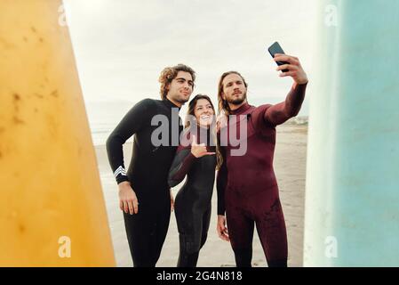 Group of happy surfer friends dressed in wetsuits standing near surfboards while taking selfie with smartphone on the beach during training Stock Photo