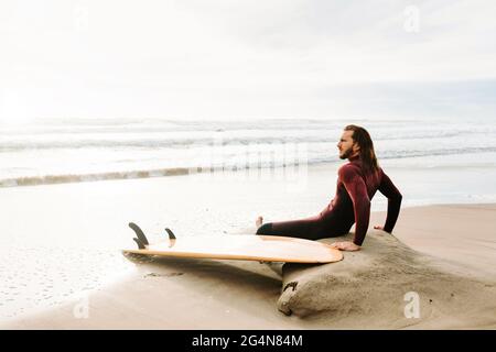 Side view of thoughtful surfer man dressed in wetsuit sitting with surfboard on the beach during sunrise Stock Photo