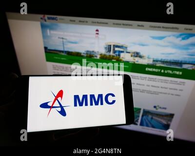 Person holding cellphone with logo of Malaysian company MMC Corporation Berhad on screen in front of business webpage. Focus on phone display. Stock Photo