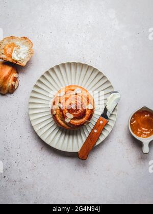 Top view of tasty cinnamon bun with almond flakes and knife with cream placed on plate near bowl with jam and napkin Stock Photo