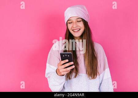 Long haired blond Hispanic female model wearing dark leather jacket and cap while smiling and looking at camera on pink backdrop in studio Stock Photo