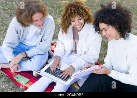 High angle of multiethnic man and women with curly hair sitting on lawn in park using laptop and sharing notepad Stock Photo