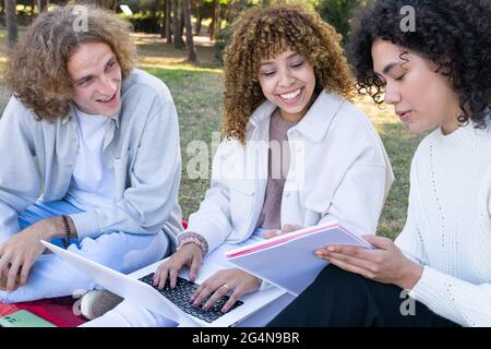 Multiethnic man and women with curly hair sitting on lawn in park using laptop and sharing notepad Stock Photo