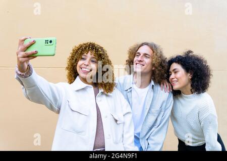 Group of young diverse women and man with curly hair using phone to take selfie together against beige wall Stock Photo