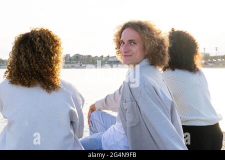 Smiling man chilling with female friends on city embankment and looking over shoulder at camera Stock Photo