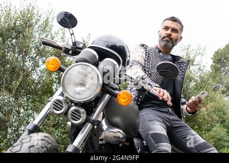 Low angle of concentrated adult brutal ethnic male biker with beard in leather jacket with rivets sitting on modern motorbike and looking away while m Stock Photo