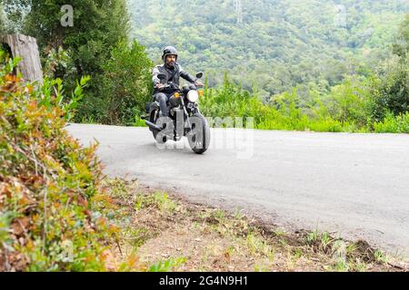 Full body of focused bearded ethnic male biker in black leather jacket and helmet riding modern motorbike on asphalt road amidst lush green trees grow Stock Photo