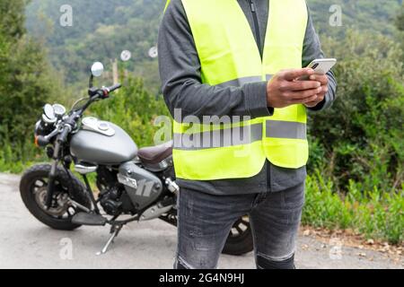Crop male biker in vest messaging on mobile phone while standing near broken motorbike near lush green woods Stock Photo