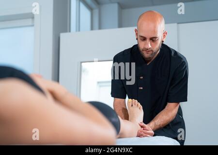 Bearded male doctor examining foot of crop anonymous Woman on table during physiotherapy in clinic Stock Photo