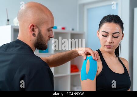Bearded male physiotherapist applying kinesio tapes on shoulder of female athlete during rehabilitation process in hospital Stock Photo