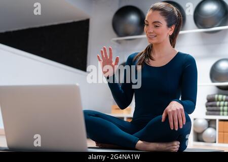 Full body of positive young female instructor waving hand while greeting students via video chat on laptop during online yoga class Stock Photo