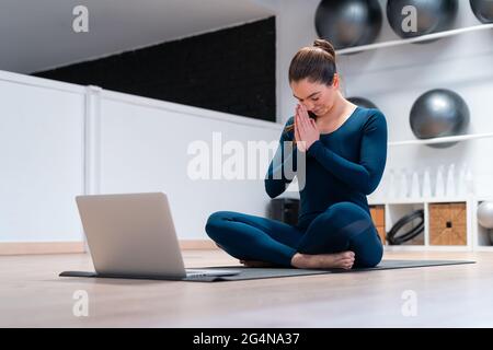 Full body of female instructor meditating with namaste hands and eyes closed while practicing yoga via video chat on laptop during online yoga class i Stock Photo