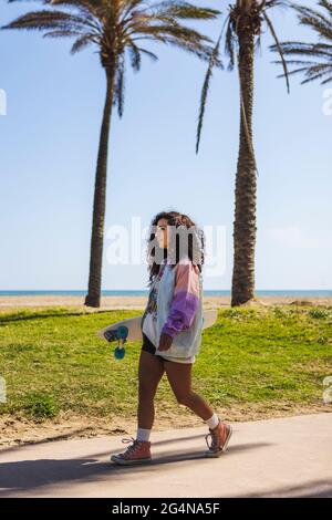 Full body side view of female with skateboard in hand strolling on sidewalk along tall palms against coast and sea Stock Photo