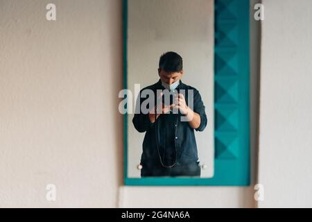 Mirror hanging on wall and reflecting man in casual clothes and mask taking photo Stock Photo